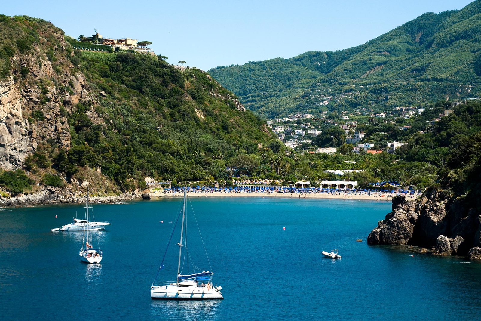 La baia a mezzaluna di San Montano vicino a Lacco Ameno, acque turchesi con barche a vela alla fonda e la spiaggia sullo sfondo